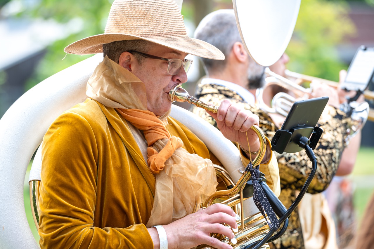 A musician in gold metallic clothes and scarves in a sunhat playing into a white sousaphone